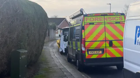 A police forensics van parked up on the side of a residential street. There is another van in front of it, with a white forensics suit hanging out of it. Police tape can be seen across the street in the distance.