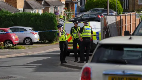 PA Media Uniformed police officers are standing on a pavement beside a road. There is blue and white police tap cordoning off a road and terraced houses are in the background.