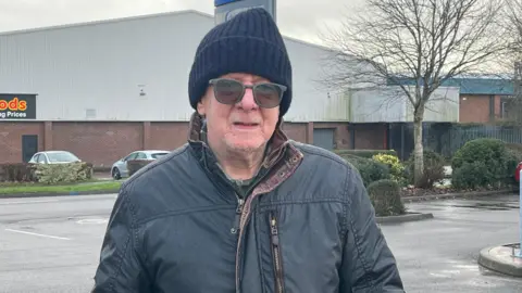 An older man with a dark blue beanie on, dark green tinted glasses and a dark navy coat. He is standing in a car park