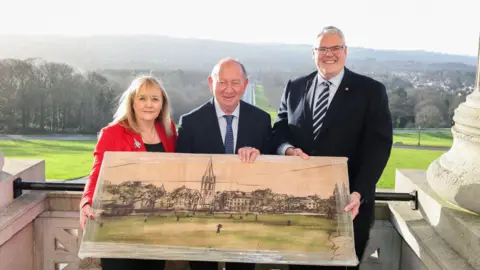 DUP A woman and two men holding a picture. The woman has short blonde hair, a black top and red blazer. The man in the middle is bald with hair on the side, he is wearing a suit and white shirt with a tie. The man at the end of the row is wearing a black blazer, patterned tie and shirt. He also has glasses.