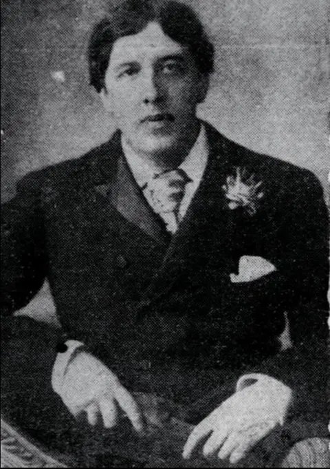 In a black and white photograph the famous poet, author and playwright Oscar Wilde is looking slightly off to the right of the camera. He's wearing a flamboyant suit with a handkerchief poking out of his top of his jacket pocket with what appears to be a a brooch pinned just above it.