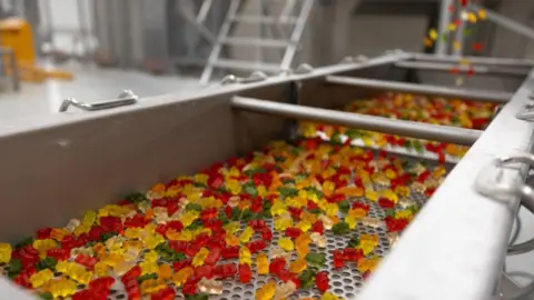 Haribo A close-up of many gummy bear sweets scattered across a white background. The bears are translucent and come in bright colours including red, orange, yellow, green, and clear, overlapping to create a colourful, textured pattern.