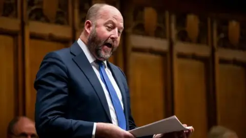 House of Commons Conservative MP Nick Timothy stands in the House of Commons. He wears a beard and a blue suit and tie with a white shirt. He is holding a sheaf of white paper. 