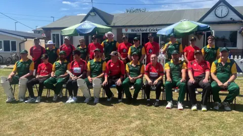 People are sat on a row of chairs with people stood behind them. There are two teams, one team is wearing a red and black uniform, and the other is wearing a yellow and green. They are outside of the Wickford Cricket Club building. 