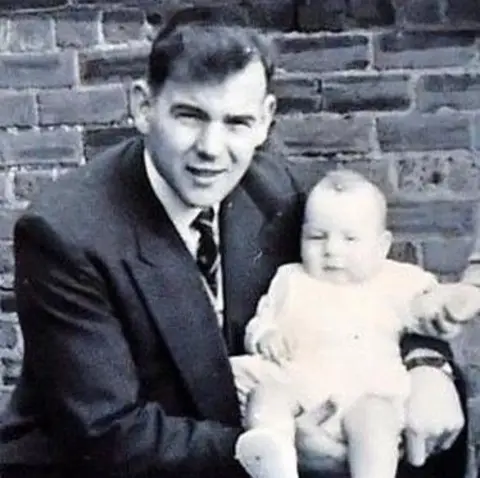 Adrian Hunt A black and white photograph of a young man in a double-breasted suit jacket, shirt and tie, crouched down holding a baby on his knee with a brick wall in the background.