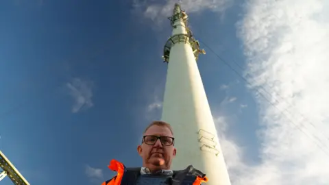 Paul Clark - a man in a bright orange high‑visibility jacket - stands at the base of a the Belmont broadcast mast, seen from a low angle. The pale mast rises sharply into a blue, lightly clouded sky, with support wires stretching outwards near the top. The angle emphasises the tower’s immense height as it looms overhead.