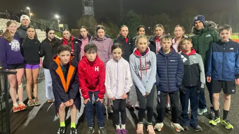 Simon Thake/BBC A large group of young athletes in sports clothes look unhappy on a wet night on the athletics track