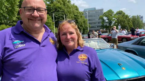 A man and a woman both wearing purple tops stand in front of a turquoise car