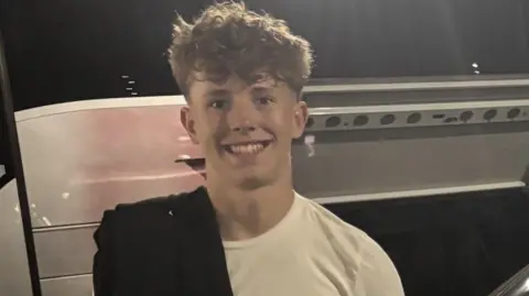 A teenage boy with curly blond hair smiles as he stands in front of a parked silver van at night. He is wearing a white T-shirt and has a black jumper chucked over one of his shoulders.