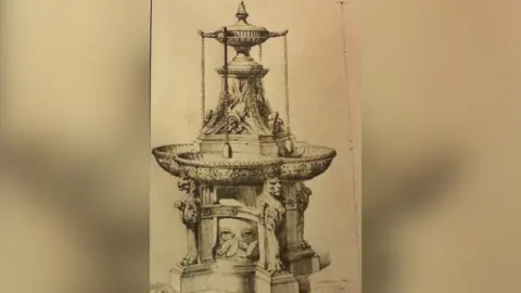 March and District Museum Black and white drawing of an ornate cast iron water feature with drinking fountain and four cups on chains hanging just above four troughs and small spouts. The structure sits on a plinth with lions heads on four supporting pillars. It stands around 2m (6ft) tall and is about 1m (3ft) in diameter.