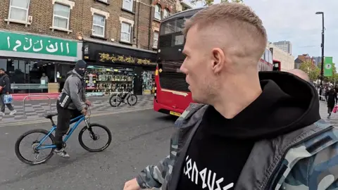 Kurt Caz A still from Kurt Caz's video in Croydon, with the YouTuber looking across the street to a cyclist who is passing by, with two shops in the background. The shop's English text has been replaced by signs which appear to be Arabic, while the cyclist has a balaclava added to his image. Kurt Caz's own image has been edited to remove the thumbs-up gesture he made to the cyclist.