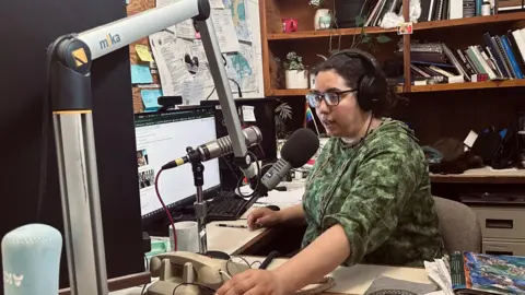 Desire Hagan, wearing a green sweatshirt, glasses and earphones, speaks into a microphone in a radio studio.
