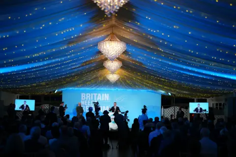 PA Media Nigel Farage on stage speaking under a chandelier during a Reform UK conference. The wide angle shot shows a line of photographers next to the stage with rows of seated audicnce members behind