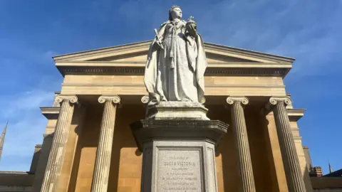 A statue of Queen Victoria holding an orb and sceptre on a plinth outside a sandstone building with tall fluted columns. Blue sky can be seen behind.