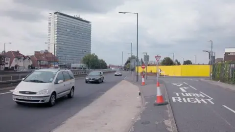 Dennis Turner via Geograph Photo taken in 2005 of a main road with cones and cars on it. There is a yellow fence to the right of the image. 