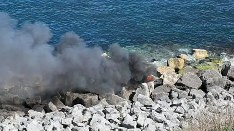 Black smoke billowing from a boat crashed onto rocks.