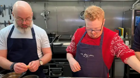 A young person with ginger hair, glasses and a red jumper wears an apron while a man next to him with a grey beard wears a blue apron. There are ovens and cooking appliances in the background. 