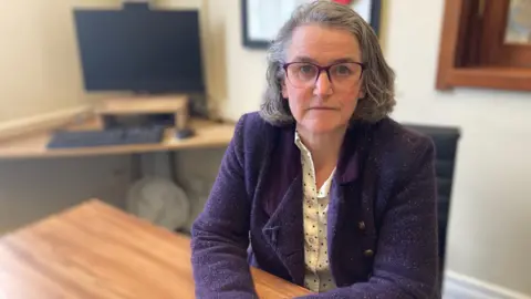 A woman sits at a desk in an office, with a computer and framed items in the background.
