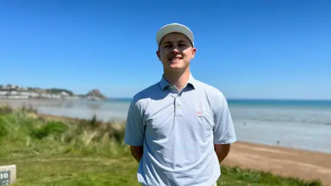 Josef smiles at the camera with his arms behind his back. There's a beach and seaside in the background along with Gorey Castle.