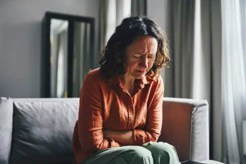 Stock image of woman with curly brown shoulder length hair sitting on a grey softa. Her arms are folded over her abdomen and she appears to be grimacing and upset in pain. She wears a terracotta shirt and green trousers. There is light coming in through a nearby window. 