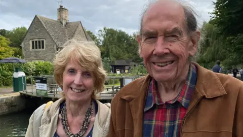 English family Judith and Sir Terence stand posing in front of a canal. Both are smiling at the camera. 