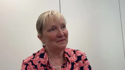 Tricia Warwick is wearing a pink and black shirt. She has blonde hair and is wearing a necklace.