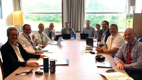 A group of people all sat around a long rectangular table, with notebooks or laptops in front of them. There are two large windows at the back of the room with greenery visible through them.