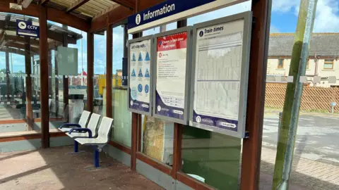 A glass shelter at a railway station contains several mounted information boards, including train times and service updates under a blue sign. A CCTV sign is visible on the left. Inside the shelter are white metal seats and a partially visible seated passenger. Outside, a road, buildings, and a wooden fence can be seen through the glass panels under a bright sky.