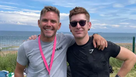 Two men, standing with their arms around each other's shoulders. The man on the left has short brown hair and he is wearing a grey t-shirt and a pink lanyard. The man on the right has short brown hair and he is wearing a black t-shirt and sunglasses. They are standing on a patch of grass and the beach and see is visible behind them.