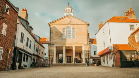 Old building in the centre of a market square with shops around the outside.