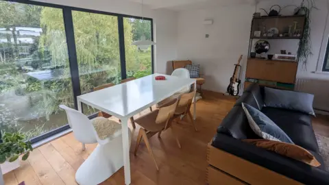 A white dining table with 60s inspired chairs and sofa. There's a guitar in the corner and a huge window showing their wildlife garden outside.