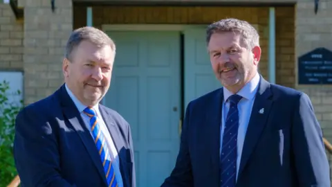 Lincolnshire County Council Sean Matthews (left) and Richard Wright, both wearing navy blue suits, standing outside a brick building in front of a pale blue double door. Metal handrails lead up to the door, and shrubs and plants surround the entrance.