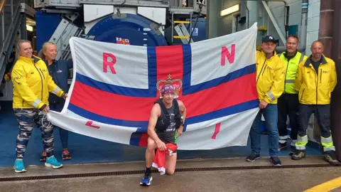 Geoff kneels down in front of an RNLI flag at Peel lifeboats station, members of the lifeboat stand on either side of the flag smile wearing bright yellow RNLI coats.