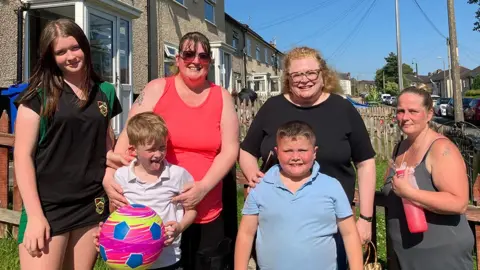 A group shot of three women, a teenage girl and two young boys standing in a garden in the sunshine