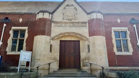 North Northamptonshire Council The outside of a building, with stone around the windows and doorway, and red brick. It has a large wooden door, with railings up to it. A sign is to the left, and it has a light slate roof. 