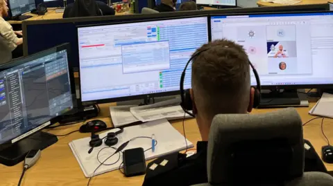 A man is sitting in an office with his back to the camera. He has three computer screens in front of him, each showing different information. He has short cropped brown hair and is wearing a dark shirt with epaulettes with pips on them. He is wearing a headset.