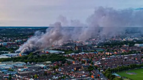 Anthony King Smoke flume starting from ground level on the left and spreading out as it goes up towards the right. The buildings of Northampton can be seen below, some with lights on as the daylight is fading