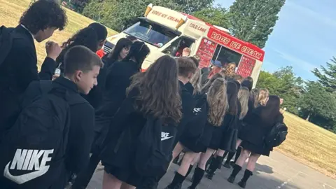 Sarah Cardwell Pupils queue up for ice cream from a van in the school grounds. The van is yellow and red with Rod's Ice Cream visible on the side of the vehicle. Pupils are dressed in black.