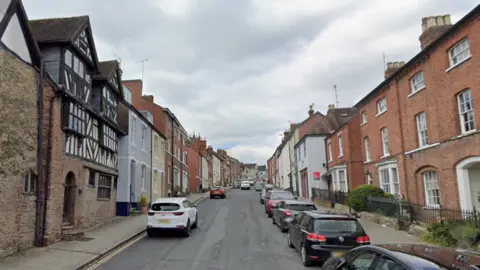 A street with rows of houses on both sides, one of them with timber frames and cars parked on both sides