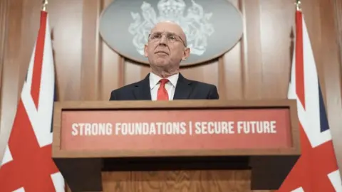 John Healey is stood behind a lectern which has the phrase "strong foundations, secure future" written on it. He is stood next to two union flags and is wearing a suit and glasses.