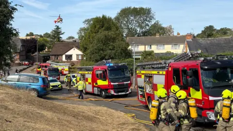 Donna Torres Several fire engines and two police cars on a street with hoses lying on the ground. Fire officers are wearing yellow helmets and have cylinders on their backs in the foreground. There is a helicopter in the sky overhead the houses in the background.