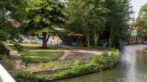 Newbury Peace Garden sits alongside a canal. It has grass trees and pathways - a number of blue barriers can be seen next to a building in the background.
