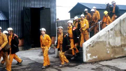 Coal miners working at Markham Colliery near Staveley, Derbyshire in 1982.  A group of 14 men wearing orange overalls and white hard-hats can be seen in the picture walking. 