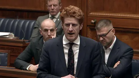 NI Assembly A man with short ginger hair wears a navy suit and white shirt with a navy floral tie. Behind him sit three men in suits. They sit at blue-leather wooden seats. 
