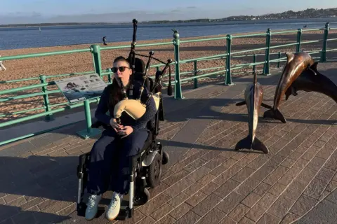 Katie Robertson A bagpiper in an electric wheelchair on the promenade of a beach next to a dolphin statue.