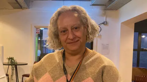 Charles Heslett/BBC A woman with a grey curly bob smiles at the camera. She is wearing a grey and pink jumper and a rainbow lanyard. She is sitting in a white room with furniture behind her.