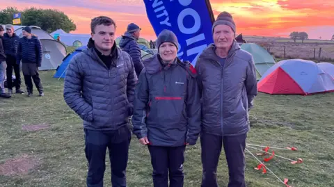 Three people standing in a line at a camp. They are all wearing outdoors gear. Tents are in the background. The sky has a pink glow as the sun is rising. 