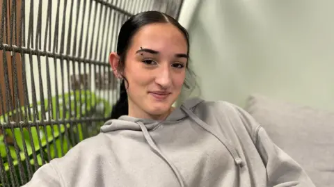 A young woman with black hair tied up in a ponytail and a pierced eyebrow wearing a light gray hoodie looks directly at the camera and smiles.