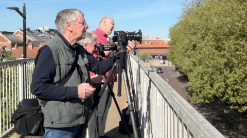 Three photographers, two men and a woman, stand on a bridge looking over metal railings. They have cameras on tripods and are looking off into the distance. To their right is a green tree and behind them are red-brick buildings. The sky is blue.