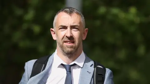 PA Daithí has white/grey short hair and a grey/ginger beard. Behind him is a blurry green background. He is wearing a black bag on his back, grey suit, white shirt and navy tie.
